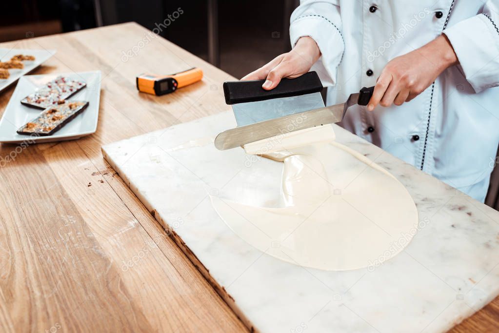 cropped view of chocolatier holding cake scrapers near melted white chocolate on marble surface 