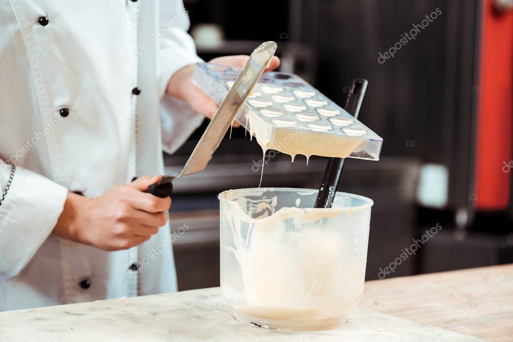 cropped view of  chocolatier pouring melted white chocolate into bowl while holding cake scraper 