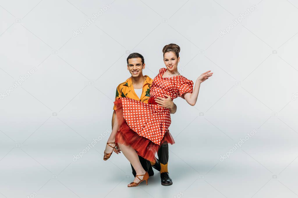 smiling dancer standing on knee and supporting partner while dancing boogie-woogie on grey background