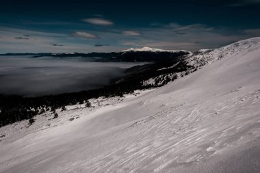 Karlı dağların manzarası, çam ağaçları ve akşam karanlığında beyaz kabarık bulutlar.
