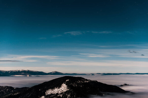 живописный вид снежных гор с соснами и белыми пушистыми облаками в темном небе вечером

