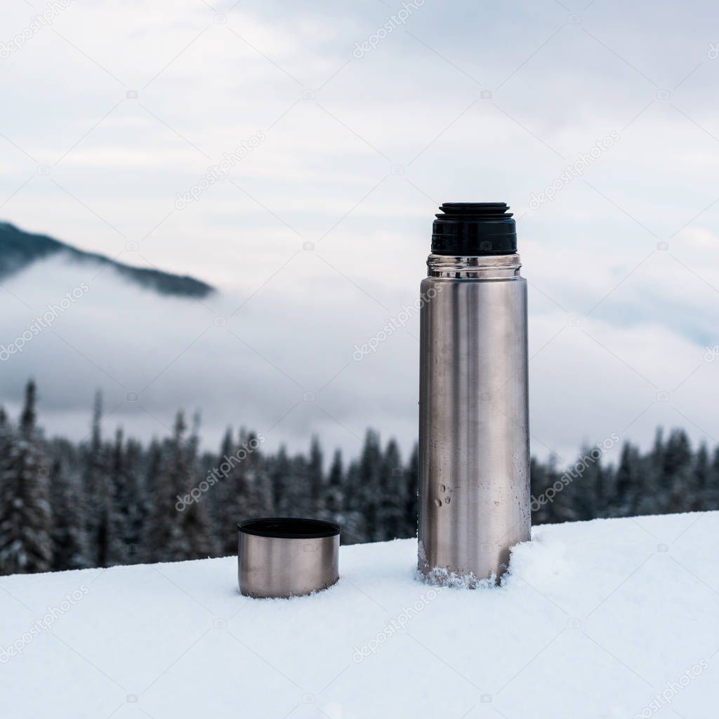 open metallic vacuum flask in snowy mountains with pine trees and white fluffy clouds