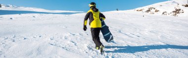 Kar üzerinde yürürken elinde snowboard tutan sporcunun panoramik görüntüsü 