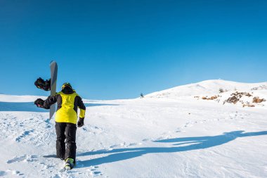 Beyaz kar üzerinde yürürken elinde snowboard tutan sporcunun arka plan görüntüsü 