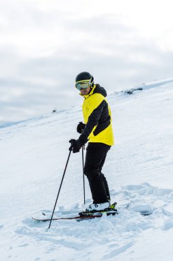 skier in helmet holding sticks and standing on slope outside  clipart