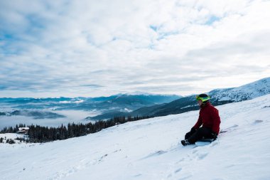 Kasklı snowboardcu mavi gökyüzüne karşı bayırda oturuyor. 