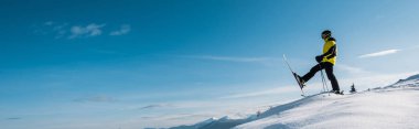 panoramic shot of skier holding ski sticks and making step against blue sky in mountains  clipart