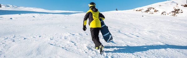 panoramic shot of sportsman holding snowboard while walking on snow 