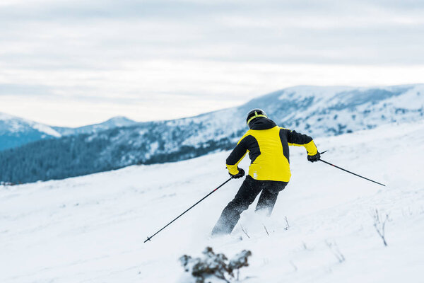 back view of skier in helmet holding sticks and skiing on slope in wintertime 