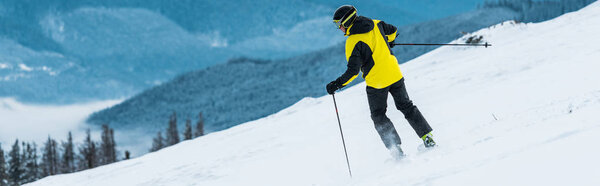 panoramic shot of skier in helmet holding sticks and skiing on slope in mountains 