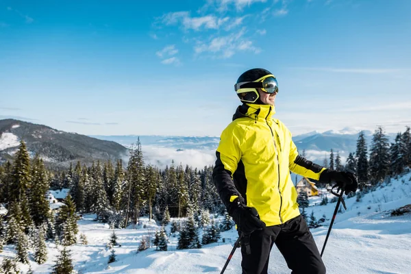 happy skier in goggles holding ski sticks against blue sky in mountains