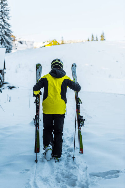 back view of skier in helmet holding ski sticks while walking on snow in winter 