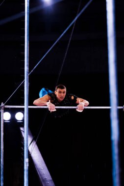 selective focus of strong gymnast performing on horizontal bars in arena of circus   clipart