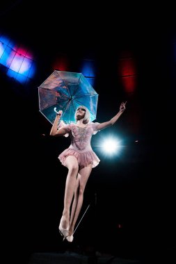 low angle view of happy aerial acrobat in costume holding umbrella and standing on rope  clipart