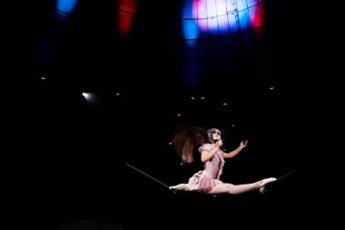 low angle view of aerial acrobat in costume holding umbrella and doing splits on rope  clipart