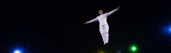 panoramic shot of handsome air acrobat with outstretched hands in arena of circus 