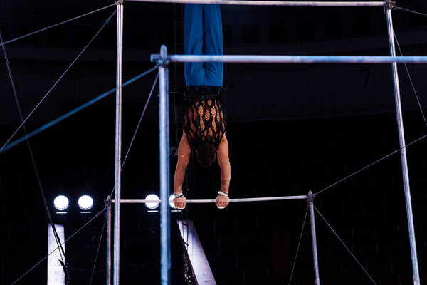 back view of athletic gymnast performing on horizontal bars in arena of circus  