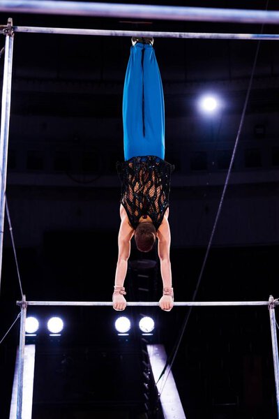 back view of strong gymnast performing on horizontal bars in arena of circus  