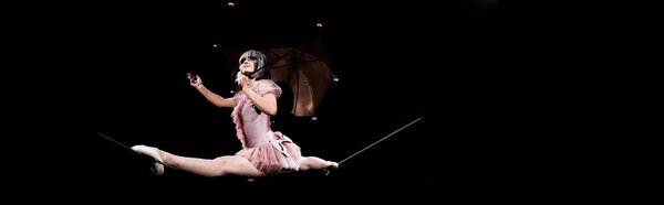 panoramic shot of aerial acrobat in costume holding umbrella and doing splits on rope 