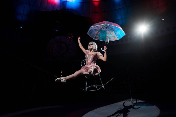 aerial acrobat holding umbrella and balancing on rope while sitting on chair 