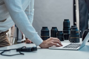 Masadaki fotoğraf lenslerinin yanında dizüstü bilgisayar kullanan sanat yönetmeni görüntüsü. 