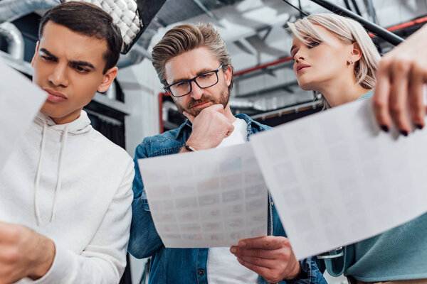 низкий угол зрения задумчивого креативного директора в очках, смотрящего на фото рядом с коллегами
 