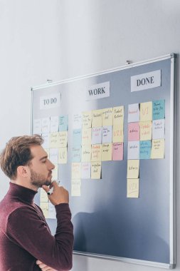 side view of handsome scrum master thinking near board with sticky notes and letters  clipart