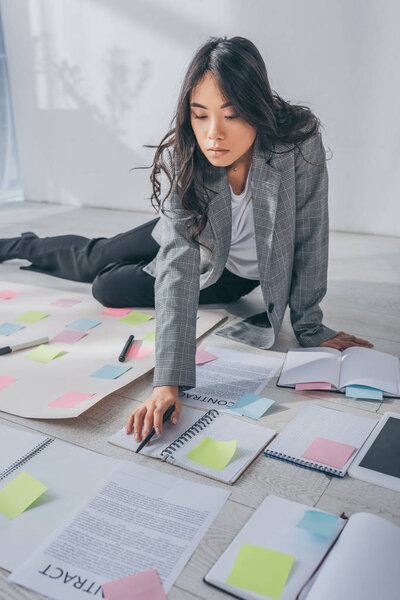 attractive asian scrum master sitting on floor and holding pen near contract 