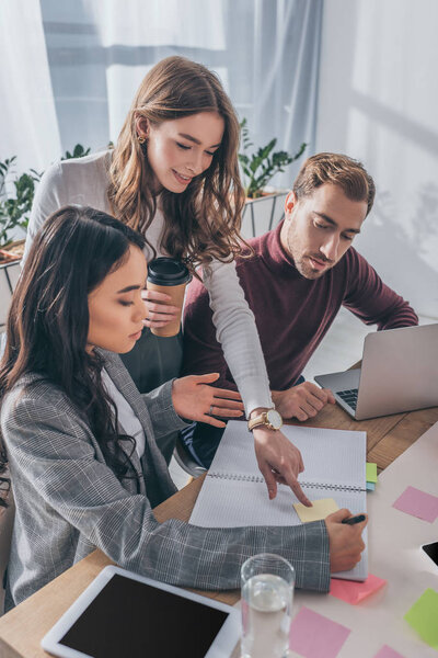 attractive businesswoman holding paper cup and pointing with finger near multicultural coworkers 