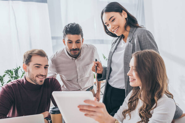 cheerful multicultural coworkers looking at digital tablet in office 