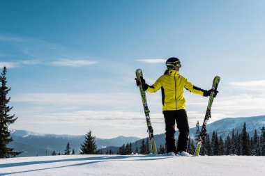 skier holding skis and standing against sky in mountains clipart