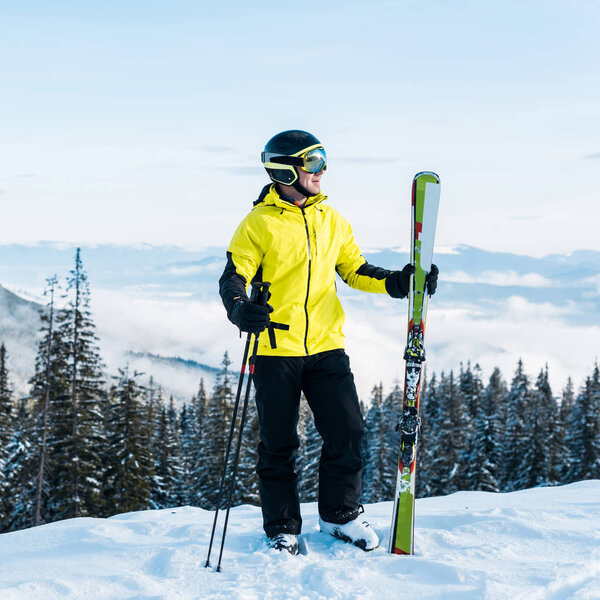 happy skier in helmet holding ski sticks and standing on snow against blue sky