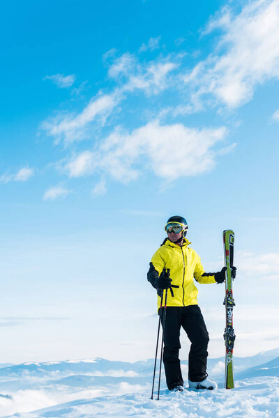  skier in goggles holding ski sticks and standing on snow against blue sky