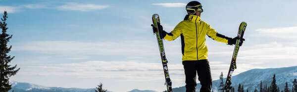 panoramic shot of skier in goggles holding ski sticks against blue sky with clouds