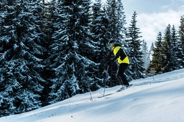 Esquiador con gafas y casco esquiando en la nieve y sosteniendo bastones de esquí cerca de los árboles —  Fotos de Stock