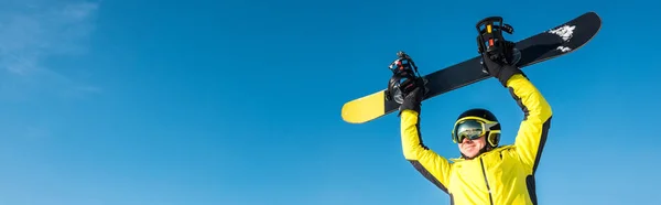 Panoramic shot of happy sportsman in helmet holding snowboard above head — Stock Photo, Image