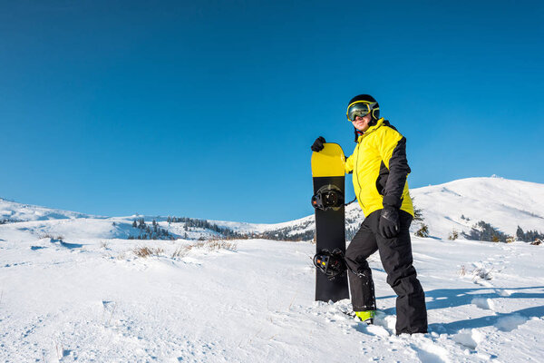 happy sportsman in helmet holding snowboard and standing on white snow in mountains