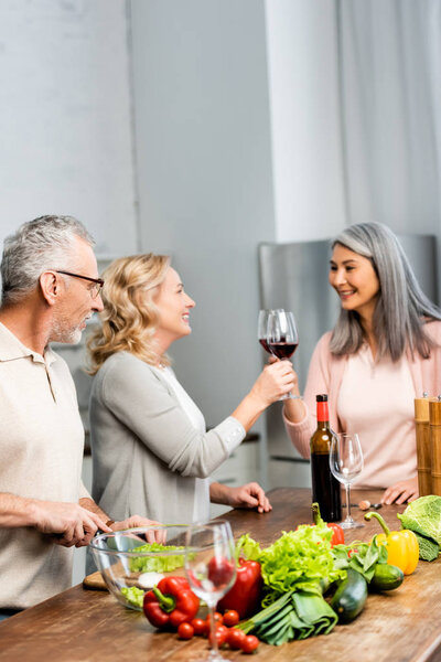 smiling multicultural woman clinking and man cutting lettuce in kitchen 