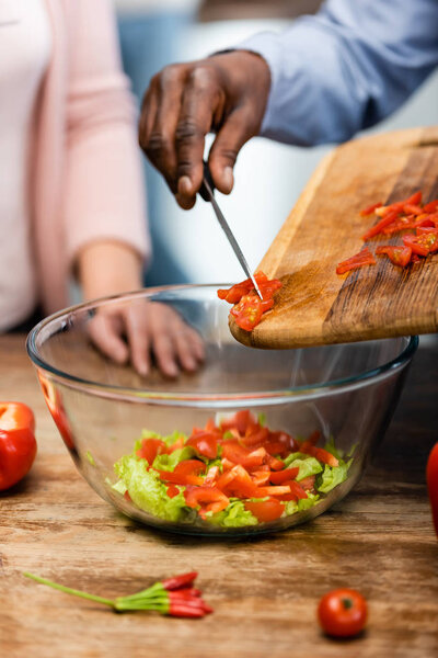 cropped view of african american man adding cut bell pepper to bowl and woman standing near him 