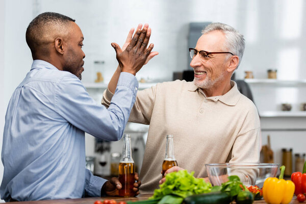 african american man giving high five to smiling friend with beer in kitchen 