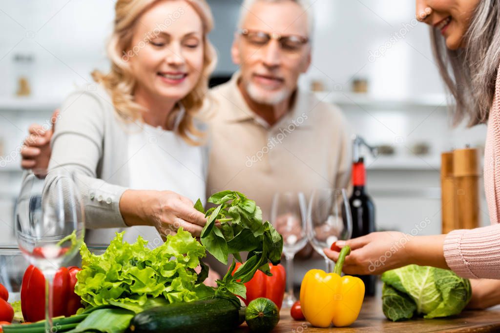 selective focus of smiling friends looking at basil in kitchen 