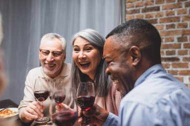 smiling multicultural friends talking and holding wine glasses during dinner  clipart