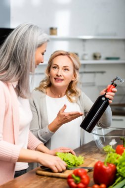 Elinde şişe, elinde tirbuşonla gülümseyen kadın ve Asain arkadaşına bakıyor. 