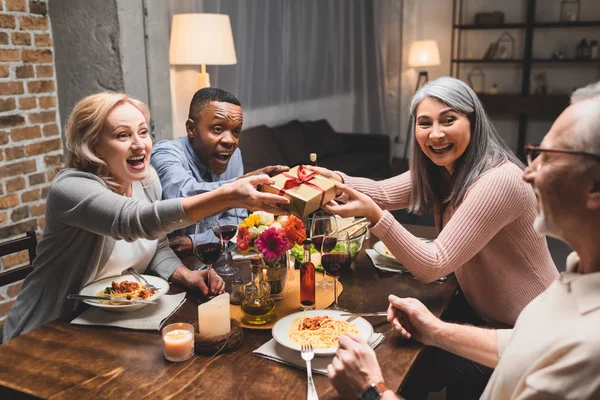 Sonrientes Amigos Multiculturales Sosteniendo Regalo Mirando Hombre Durante Cena —  Fotos de Stock