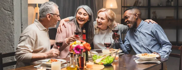 夕食中にワイングラスを抱いたり抱き合ったりする多文化の友人たちの笑顔のパノラマ写真 — ストック写真