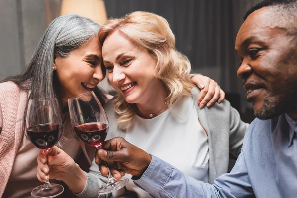 smiling multicultural friends hugging and clinking with wine glasses during dinner