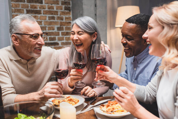 улыбающиеся мультикультурные друзья разговаривают и звенят с бокалами вина во время ужина
 
