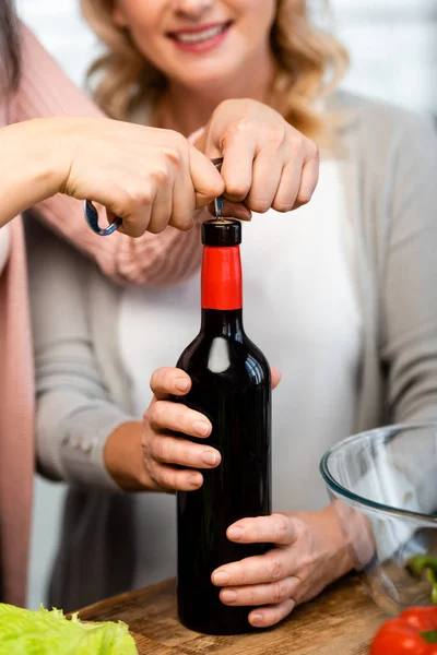 cropped view of friends opening wine bottle with corkscrew in kitchen