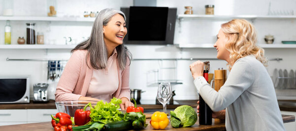 panoramic shot of smiling woman opening wine bottle with corkscrew and looking at her asian friend 