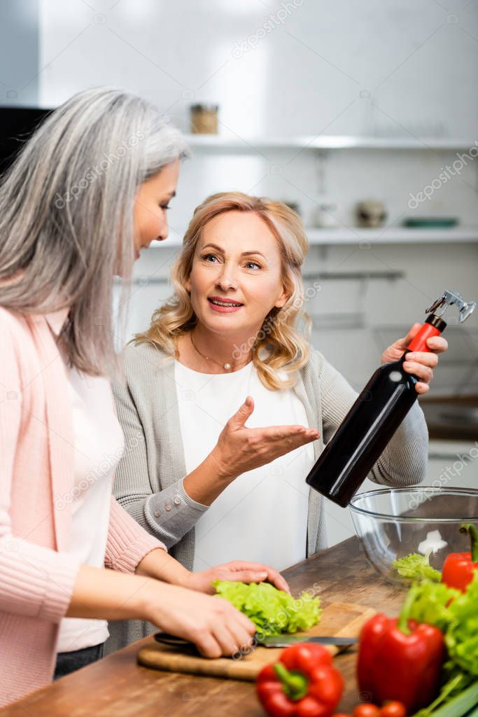 smiling woman pointing with hand at bottle with corkscrew and looking at asain friend 
